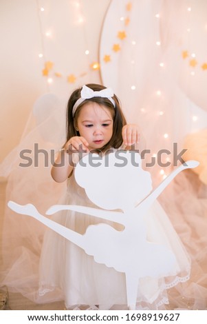 Cute little girl in a white princess dress with a bow on her hair plays with wooden ballerinas among the golden stars on the background of a large white wooden moon and garlands. Children's decor. 