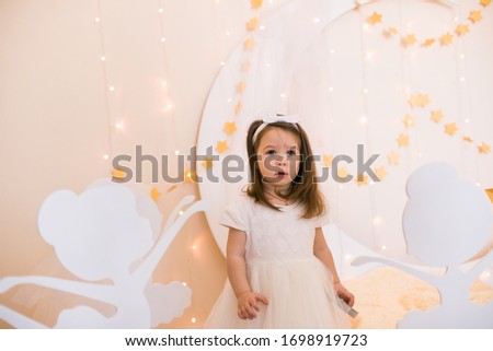 Cute little girl in a white princess dress with a bow on her hair plays with wooden ballerinas among the golden stars on the background of a large white wooden moon and garlands. Children's decor. 
