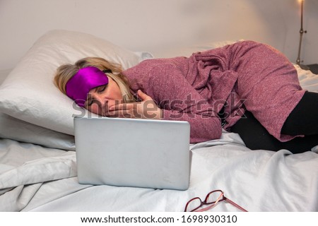 woman in bed with bathrobe and eye mask reading on her computer and yawning