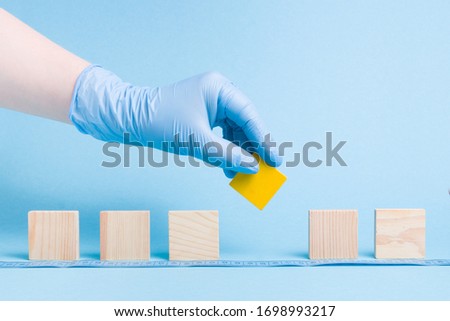 hand in rubber disposable blue medical glove takes a yellow block a wooden square, dominoes are lined up, blue background, copy space, social distance concept, isolation of a sick person from society