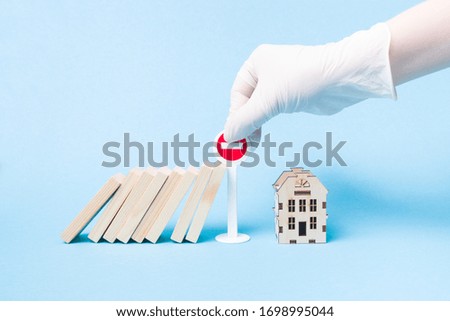 hand in a white medical rubber glove stops a domino using a miniature stop road sign, wooden house model, blue background, self-isolation and quarantine concept, stop a pandemic, stay home