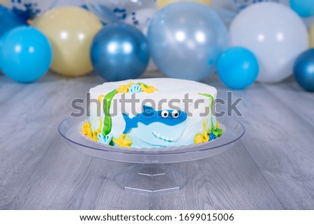Close-up shark cake decorated with multicolor buttercream icing. Birthday cake with shark motifs. Birthdays kids.White-blue-yellow and glittering balloon background.Bakery. Baby shower party.