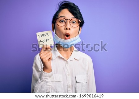 Young beautiful asian girl wearing medical mask holding reminder with virus alert message scared in shock with a surprise face, afraid and excited with fear expression