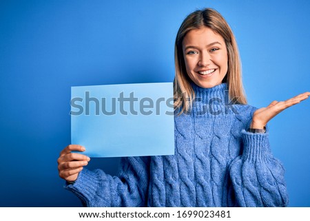 Young beautiful woman holding banner standing over isolated blue background very happy and excited, winner expression celebrating victory screaming with big smile and raised hands