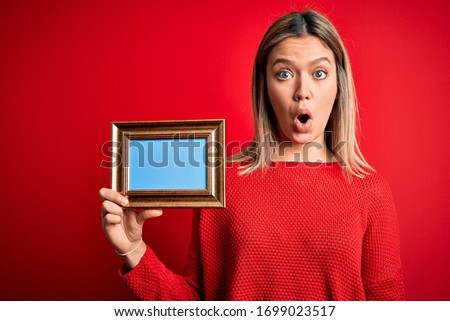 Young beautiful woman holding vintage frame standing over isolated red background scared in shock with a surprise face, afraid and excited with fear expression