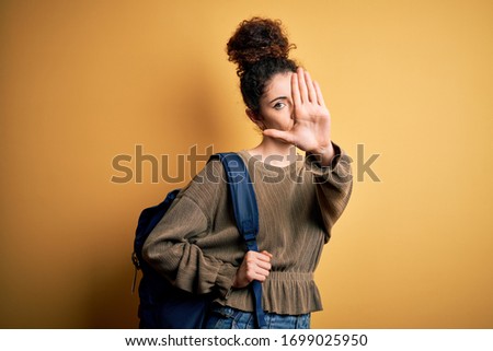 Young beautiful student woman with curly hair and piercing wearing backpack with open hand doing stop sign with serious and confident expression, defense gesture