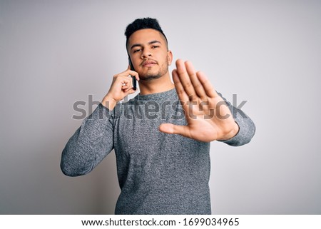 Young handsome man having conversation talking on the smartphone over white background with open hand doing stop sign with serious and confident expression, defense gesture