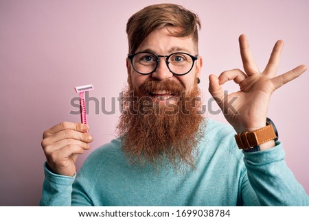 Redhead Irish man with beard holding beauty razor for shaving and skin care over pink background doing ok sign with fingers, excellent symbol