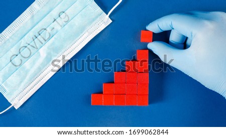 Hand in glove adding red cubes to growing column graph representing a rising number of infected or death toll of Corona Virus pandemic. Royalty-Free Stock Photo #1699062844