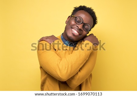 Pleased african american man in yellow clothes and glasses hugs himself, has high self esteem. Studio shot on colored wall. Royalty-Free Stock Photo #1699070113