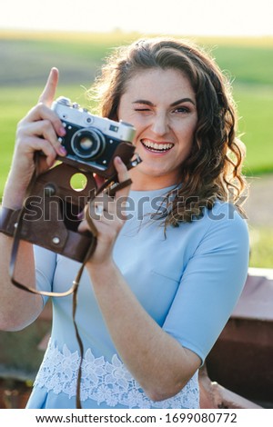 Close Up portrait of smiling beautiful girl taking photo on retro camera. A bright girl in a blue dress is smiling and holding a retro camera in her hands. Girl with a camera at the cottage