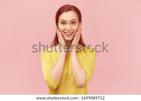 Happy woman touches cheeks, expresses gratitude, smile laughs very happy, surprised by an unexpected gift. Young girl with long red hair dressed in sweater stands isolated on pink background in Studio