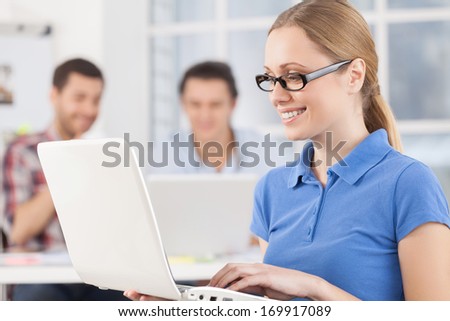 Happy to be a part of the team. Cheerful young woman working on laptop and smiling while her colleagues working on the background