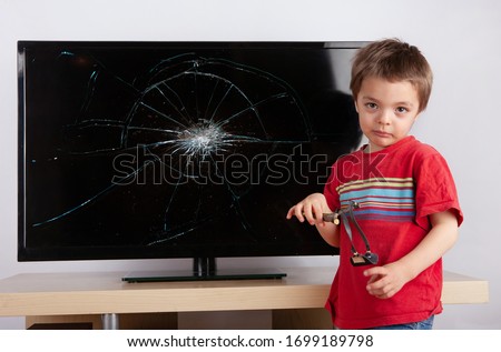 Cute little boy standing in front of a TV with broken screen with his slingshot.