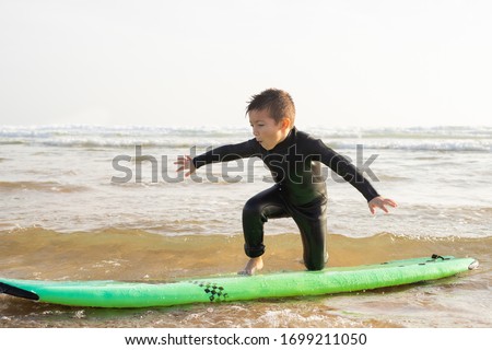 Cute little boy learning to be surfer. Talented child in swimwear training his balance on green surfboard with sea waves on background. Vacation, surfing and summer concept