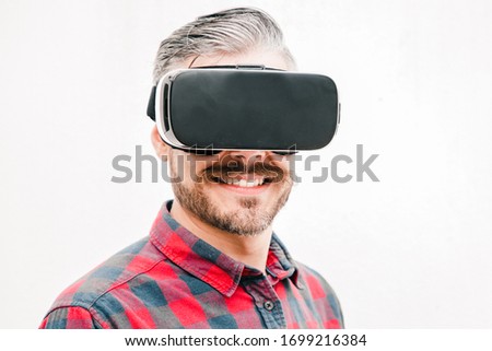 Close-up view of content man in VR headset. Handsome happy bearded man in checkered shirt using virtual reality headset on grey background. Technology concept