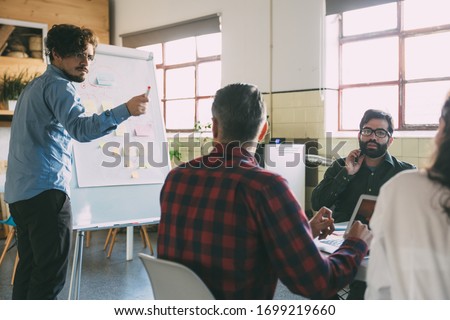 Creative team discussing ideas for startup project, speaker at board pointing at audience. Business colleagues in casual working together in contemporary office space. Brainstorming concept