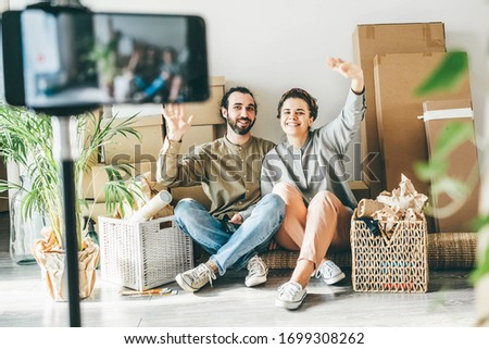 Lovely joyful young couple with belongings packed in different boxes sits on floor filming video with mobile phone in new apartment on moving day.