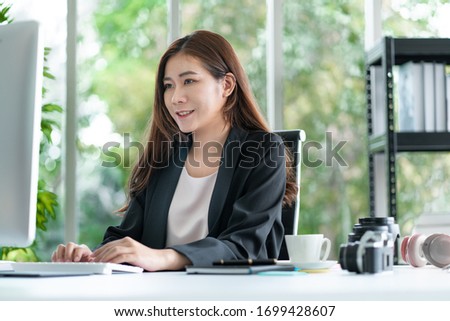 Asian business woman sitting at her work desk looking at computer monitor with smile while camera and lens on her desk. Photography business concept.