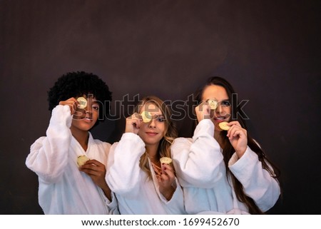 Session in a beauty salon. Three happy girlfriends with cucumbers on their eyes. Blonde girl, brunette girl and girl of Caribbean American origin with afro hair
