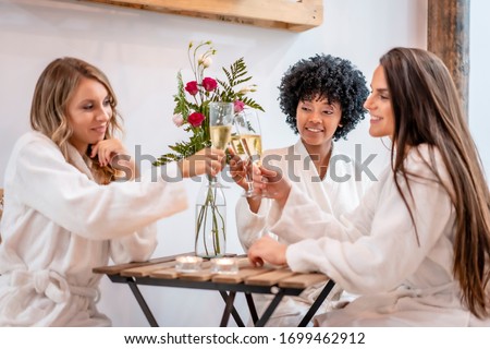 Session in beauty salon. Three young models in white coats sitting after the treatment toasting with champagne. Blonde girl, brunette girl and girl of Caribbean American origin with afro hair
