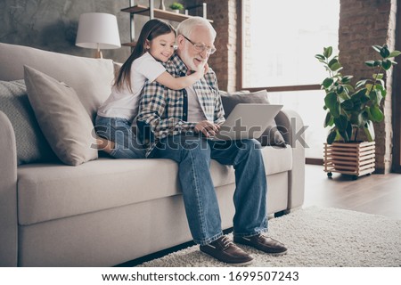 Profile photo of funny old grandpa little granddaughter sit sofa stay house quarantine safety watch movie notebook talk skype relatives modern design interior living room indoors