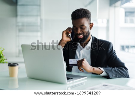African man in casual outfit talking on phone and reading credit card number while sitting at office