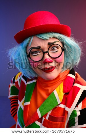 Close-up portrait of a clown girl with make-up, blue hair, a red hat, a colored checkered jacket and glasses. A clown shows different human emotions. April Fools Day concept. 