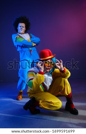 Two clowns a man and a woman with makeup in colored tracksuits in the style of the 80s or 90s posing against a trendy neon background.Party disco 80s 90s vibes.Creative art purple color style