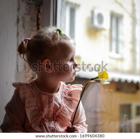 A cute girl sits at the window during quarantine. The child holds a yellow flower in his hands. Beautiful baby is bored at home on the windowsill.
