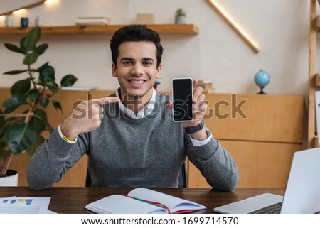 Businessman looking at camera, smiling and pointing at smartphone at table in office