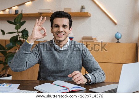 Businessman looking at camera, smiling and showing okay sign at table in office