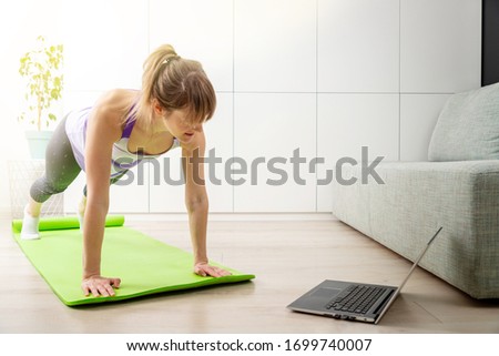 Girl makes a bar on a fitness mat at home in the living room looking at a workout (exercise) online on a laptop. Copy space.