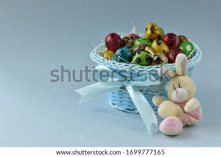 The rabbit sits cross-legged at the basket with Easter quail eggs. On a blue background is a blue basket with Easter eggs.