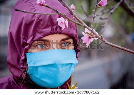 A girl with beautiful colour of eyes in the blue mask and in the pink protective suit protects herself from a pandemic, viruses, coronavirus against the background of the blossoming tree in spring.