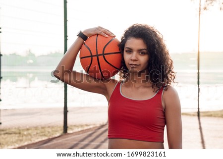 Young woman basketball player standing holding ball outdoors on court posing to camera smiling joyful close-up