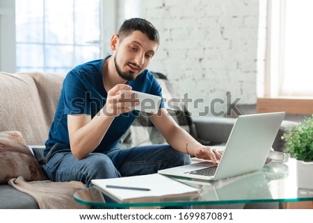 Young focused man studying at home during online courses or free information by hisself. Becomes economist, financist, translator while isolated, quarantined. Using laptop, smartphone, headphones.