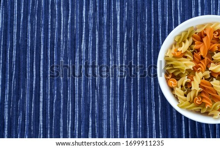 Raw colored spiral pasta in a white bowl on a striped white blue cloth background with a side. Close-up with the top. With space for text.