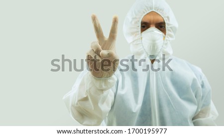 Doctor with mask and bioprotective suit with arm extended forward and his right hand making a sign of peace and love on white background