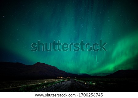 Massive northern lights over an empty gravel road in Iceland-. The sky is clear and the stars are bright. The empty gravel road leads up to a mountain.