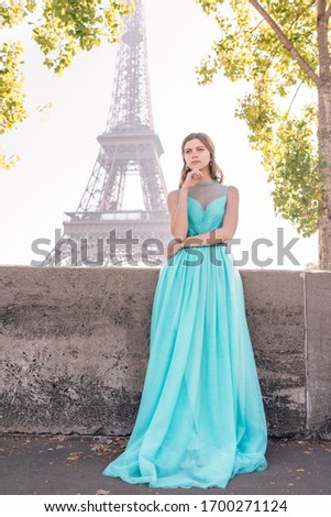 A young beautiful and elegant girl in a blue and green dress stands against the background of the Eiffel tower in Paris.