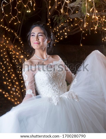 Teenager Latina Mexican posing with a white quinceañera dress and white roses in a garden at night, with a tree on the background surrounded with small warm lights.