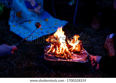 Two campers grilling miniature cinnamon buns on a single use aluminum camping grill. The fire burns strong. Another camper wearing a blue blanket with stars is visible in the background.
