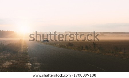 Picture of long road goes far away to horizon in middle of countryside. Tractor and car with people standing besides road. Sunrise or sunset time. Harvest period