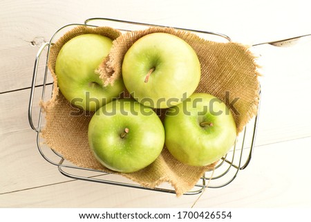 A group of several organic juicy, ripe, sweet, bright green apples with a wicker basket of wire and a jute napkin, close-up, on a background of natural wood.