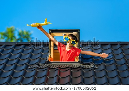 The boy plays with a toy airplane on the roof of the house. Blue summer sky on background. The concept of successful start and business. The roof is covered with metal tiles. Happy summer vacation.