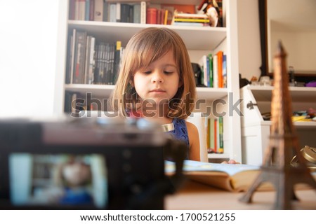 Trendy little girl in summer casual wear and white hat is recording herself with a camera at home. On the desk there is a metal souvenir of Eiffel tower. White library on background. Little blogger.