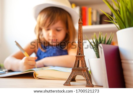 Trendy little girl in summer casual wear and white hat, is doing  homework or writing notes on map. In foreground a metal souvenir of Eiffel tower, succulents and passports. Library on background.