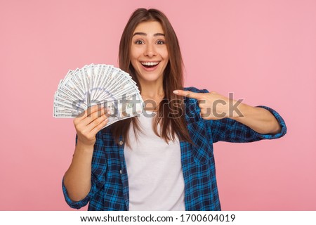 Portrait of joyful lucky girl in casual shirt happy to hold lot of money, pointing at dollar bills in hand and smiling excitedly, enjoying lottery win. indoor studio shot isolated on pink background Royalty-Free Stock Photo #1700604019