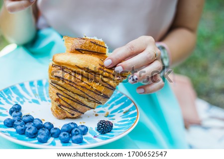 young girl's hand holding a croissant. croissant with blueberries on a blue plate. young girl eating a croissant in a backyard. closeup of a fresh croissant in hands. breakfast ideas in isolation